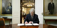 Der Finnische Präsident sitzt an einem glänzenden Holztisch, hinter ihm Gemälde "wichtiger" Männer und und unterschreibt Papiere