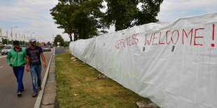 Zwei Flüchtlinge laufen an einem Zaun vorbei, an dem „Refugees welcome“ steht