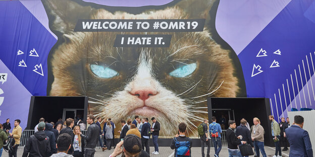 Menschen stehen vor einer Messehalle mit einem Konterfei einer Katze und der Aufschrift "Welcome to #OMR19 I hate it."