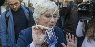 Clara Ponsati, eine Frau mit weißen Haaren, hält einen Ausweis in die Höhe