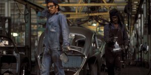Zwei Arbeiter in blauen Anzügen vor einem Auto in einer Produktionsstraße