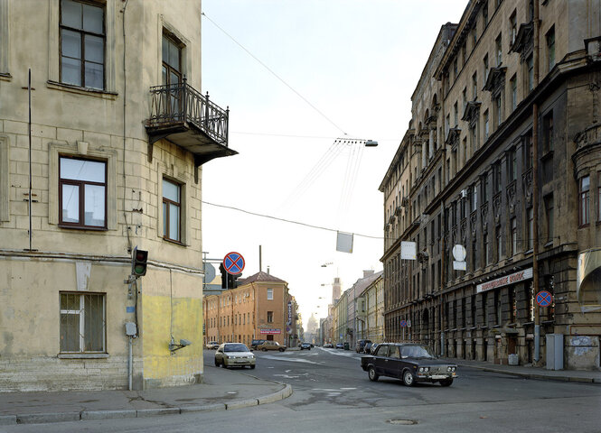 Aufnahme einer Straße in Farbe. Zu sehen ist eine Straße in Lima, die eine kurve nach links nimmt. Im Bild ganz links ist eine Häuserfassade in beigen Farbtönen zu sehen, auf der gegenüberliegenen Straßenseite ein Altbau mit dunkelgrauer Witterung an der Fassade. Ein schwarzes Auto fährt von linke nach Rechts ins Bild.