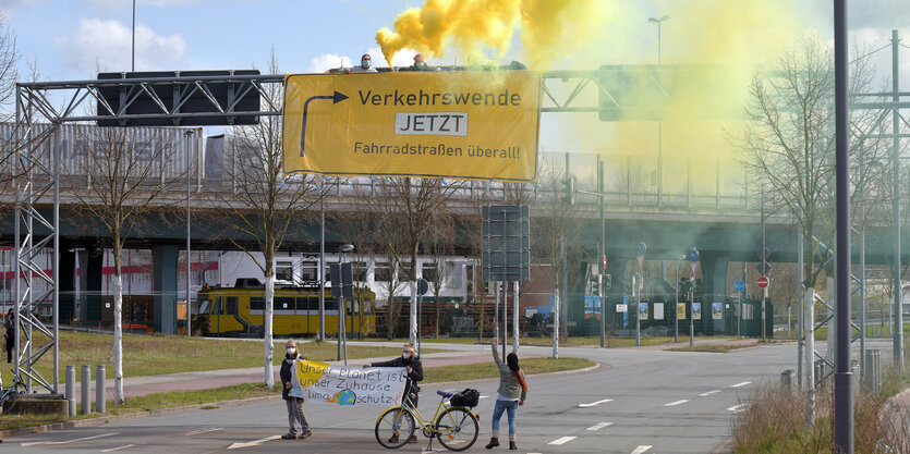 Menschen mit Fahrrädern auf einer merhspurigen Straße, über ihnen hängt ein Transparent mit der Aufschrift "Verkehrswende jetzt - Fahrradstraßen überall"