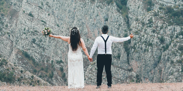 Ein Hochzeitspaar hält sich an den Händen und schaut auf beeindruckende Berg-Kulisse, Blick von hinten