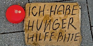 Ein Pappschild mit Aufschrift "Ich habe Hunder Hilfe Bitte" auf dem Fußweg, daneben eine rote Schale