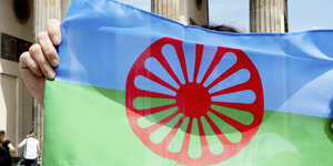Auf dem Bild ist eine Roma-Flagge zu sehen, die vor dem Brandenburger Tor hochgehalten wird. Die Person, die sie hochhält, ist nicht zu sehen. Auf die Roma-Flagge haben sich die Delegierten auf dem ersten Weltromakongress geeinigt, am 8.4.1971. Deshalb ist der 8. April der Weltromatag. Sie einigten sich außerdem auf die Selbstbezeichnung Roma und die Hymne "Gelem, gelem". Der Weltromatag wird weltweit gefeiert.