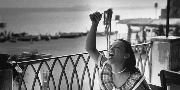 Black and white photo of a woman eating spaghetti on a seaside balcony