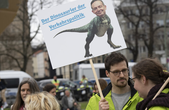 Ein Mensch trägt ein Plakat, auf dem Wissing als Dinosaurier abgebildet ist