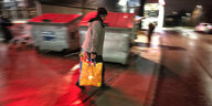 Eine Frau mit orangenen Plastiktaschen vor zwei Containern am Abend