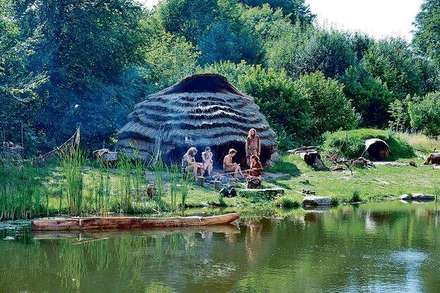 Als Steinzeitmenschen verkleidete Darsteller sitzen vor einer Hütte um ein Feuer.