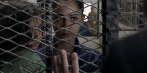Eine Frau und ein Kind schauen durch ein Gefängnisgitter, auf der anderen Seite steht ein Mensch