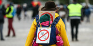 Eine Person in gelber Jacke von hinten. Sie trägt einen Hut. Auf dem Rücken in einer Kraxe ein Kleinkind, das Mütze trägt. Auf dem Rücken des Kleinkinds ein Schild, auf dem eine Spritze durchgestrichen gezeichnet ist.