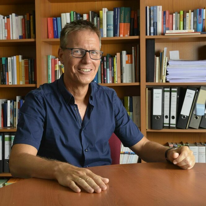 Michael Meyen, ein älterer Mann mit kurzen Haaren, sitzt in kurzärmligem Hemd an einem Schreibtisch, auf den er seine Unterarme und Hände legt. Meyen trägt Brille