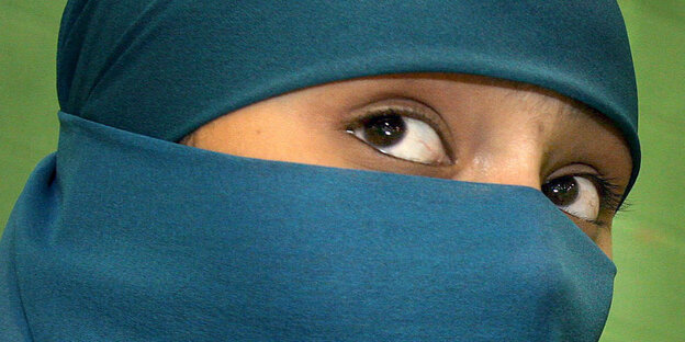 Im Bild zu sehen ist eine Frau, die einen blauen Niqab trägt, der nur ihre Augen freilässt, vor einem grünen Hintergrund.