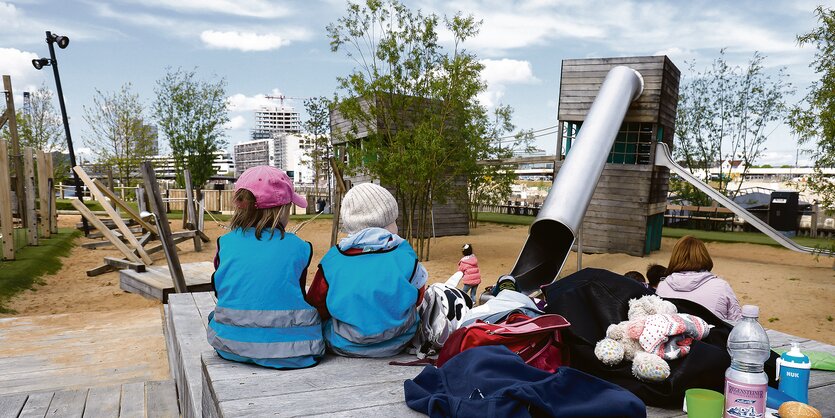 Zwei Kinder in blauen Jacken sitzen auf einem Podest, dahinter eine Sandfläche mit Rutschenturm