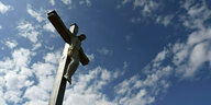 Eine Figur des Jesus hängt an einem Holzkreuz vor blauem Himmel mit Wölkchen