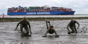 Bei der Wattolympiade vergnügen sich Leute im Schlick, während ein Containerschiff vorbeifährt.