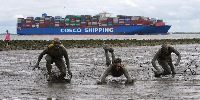 Bei der Wattolympiade vergnügen sich Leute im Schlick, während ein Containerschiff vorbeifährt.