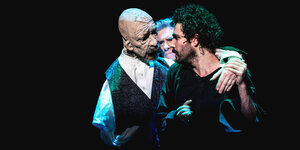 Eine Schauspielpuppe mit einem Männerkopf und zwei Männer stehen umschlungen vor einem dunklen Hintergrund