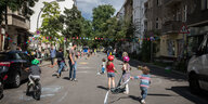 Spielende Kinder und vereinzelt Erwachsene in einer städtischen Straße. Wenige Autos parken am Straßenrand. Es ist Sommer.