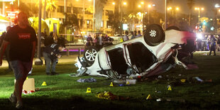 Ein überschlagendes Auto liegt auf einem Rasenstück. Die Polizei markiert Indizien, im Hintergrund eine durch Straßenlichter erleuchtete Häuserzeile. Es ist später abend