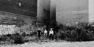Die drei Mitglieder der Band Die Letzten Ecken stehen auf einem Schwarz-Weiß-Foto, das eine Totale zeigt, im Freien. Sie stehen vor zwei Häuserwänden, die hinter ihnen zu einem Dreieck zusammenlaufen, dass einen Spalt frei lässt. Am Boden sind Grasbüschel und ein Straßenbelag aus Pflastersteinen zu sehen.