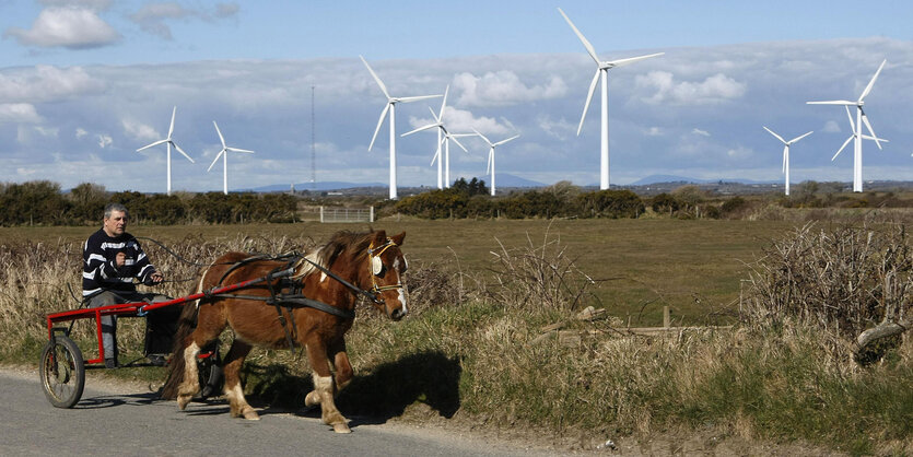 Eine Kutsche fährt auf einer kleinen Straße, im Hintergrund stehen Windräder