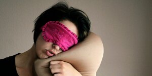 Eine junge Frau trägt eine pinkfarbige Schlafmaske und hat den Kopf auf ein Kissen gelegt