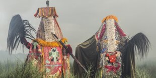 Zwei von Masken und Kleidern verhüllte Figuren stehen zwischen hohen Halmen