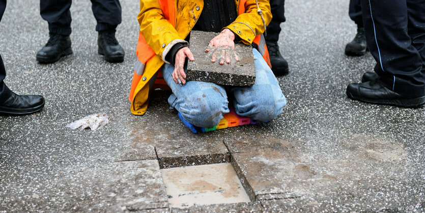 Eine Aktivistin der Letzten Generation, umstanden von Polizisten, auf ihrem Schoß liegt das aus der Straße ausgesägte Stück Asphalt, auf das sie ihre Hand geklebt hat