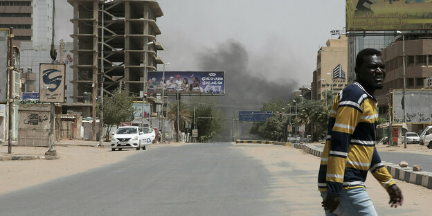 Eine Straße in Khartum. Im Hintergrund steigen Rauchwolken auf. Im Vordergrund geht ein Mann über die Straße.