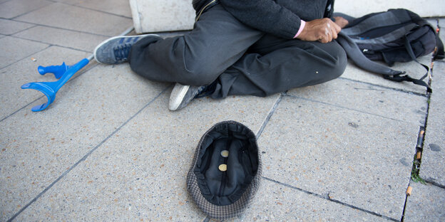 Ein Mann sitzt auf dem Boden hinter einer Mütze, in der sich Münzen befinden. Neben dem Mann liegt eine Krücke.