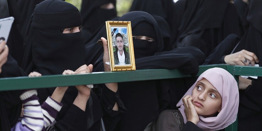 Ein Mädchen blickt auf das Portraitfoto eines Mannes, es steht in einer Menge von Frauen, die Tschadors tragen