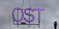 Ein lila leuchtendes Schild auf dem Ost steht, hängt an eine Metallkette in der Luft.