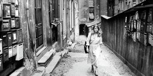Still aus Claudia von Alemanns Schwarz-weiß "Reise nach Lyon". Eine Frau im Trench Coat läuft eine Gasse in Lyon entlang, deren Wände von Briefkästen gesäumt sind. An den Wänden und am Boden gibt abgebröckelter Putz die Sicht auf Backsteine frei.