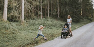 Eine Frau schiebt einen Kinderwagen auf einem Waldweg, ein Kind springt aus dem Wald