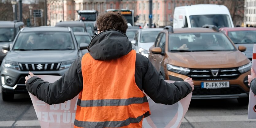 Ein Aktivist in Rückansicht beim Blockieren einer Straße, im Hintergrund wartende Autos und der Brandenburgische Landtag