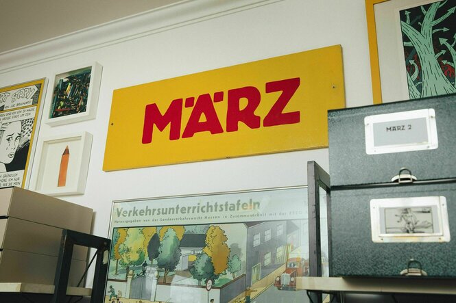 Ein Schild "März" mit der bekannten roten Schrift auf gelbem Untergrundhängt an der Wand zwischen Bildern