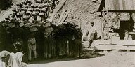 Wir sehen den Ausschnitt eines Fotos von circa 1942. KZ-Häftlinge in Reihen bewegen sich mit schweren Granitsteinen auf dem Rücken über eine breite Treppe nach oben