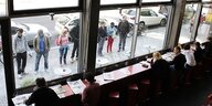 Eine Reihen von Menschen steht vor den Fenstern eines Cafés, innen sitzen Cafébesucher und -besucherinnen