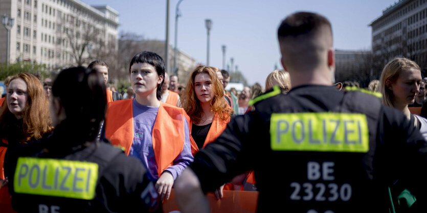 Polizist:innen von hinten. Über ihre Schultern hinweg sieht man Menschen in Warnwesten und die Prunkbauten der Karl-Marx-Allee