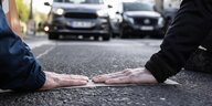 Hände von Klimaktivistinnen auf dem Asphalt einer Straße, Autos stehen den Aktivistinnen gegenüber