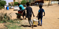 Zwei Kinder tragen einen Eimer mit Wasser an einem Straßenrand