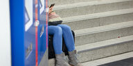 Ein Fahrgast sitzt auf einer Treppe zu den Gleisen und blickt auf sein Smartphone.