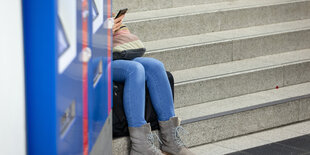 Ein Fahrgast sitzt auf einer Treppe zu den Gleisen und blickt auf sein Smartphone.