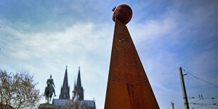 Eine Figur steht im Vordergrund, im Hintergrund ist der Kölner Dom zu sehen