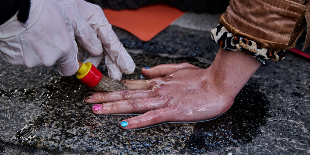 Bunt lackierte Fingernägel an einer Hand, die auf der Strasse klebt, Hände mit Handschuhen und Pinsel bearbeiten die Hand