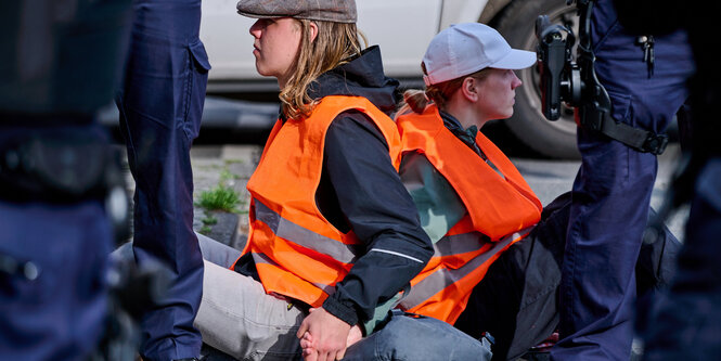 Zwischen Polizistenbeinen sitzen Blockiererinnen in gelben Schutzwesten und halten sich an den Händen