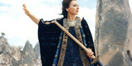Filmstill aus Pasolinis "Medea". Eine Frau in einem Gewand aus schwerem blauen Stoff steht vor einer felsigen Landschaft. Sie hält eine Schaufel in der linken Hand und hebt den rechten Arm.