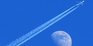 Ein Flugzeug fliegt durch strahlend blauen Himmel und hinterlässt einen Kondensstreifen quer über das gesamte Bild. Im Hintergrund ist der Mond groß zu sehen. Es sieht so aus, als würde das Flugzeug am Mond vorbeifliegen.